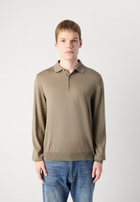 Ung mann iført beige langermet polo skjorte og blå jeans, stående mot en enkel hvit bakgrunn, vendt framover med en nøytral uttrykk.