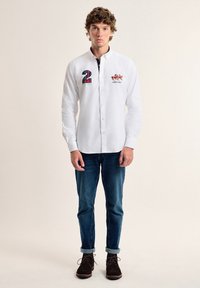Camisa de botones blanca con bordado; presenta un parche de cuadros azules con el "2" en un lado y un logo en el otro. Combinada con jeans azules y zapatos marrones.