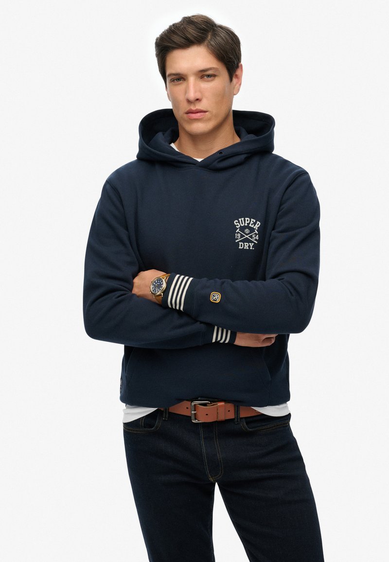 Felpa blu navy con logo bianco, polsini rigati e tasca frontale. Indossata con jeans scuri e una cintura marrone, con un orologio al polso.