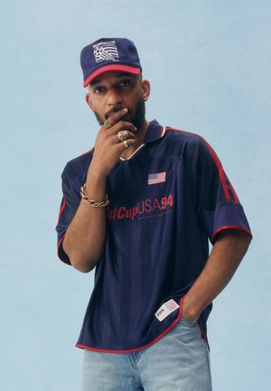 Muž oblečený v tmavomodrom tričku a šiltovke World Cup USA 94, pózujúci s rukou na brade a nosiaci niekoľko prsteňov a náramok.