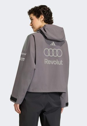 Personne portant une veste grise à capuche avec les logos des anneaux Audi, Revolut et Adidas dans le dos, debout de profil sur un fond uni.