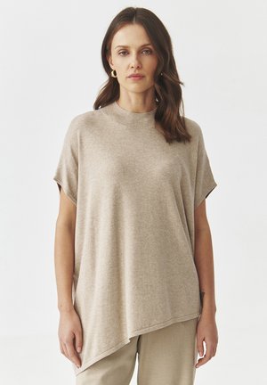 Pull beige surdimensionné avec un col montant, des manches courtes et un ourlet asymétrique. Texture douce, coupe décontractée, complète les tons neutres.