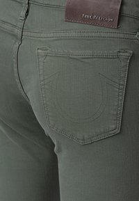 Jeans de mezclilla en verde oliva con un diseño distintivo bordado en el bolsillo trasero y una etiqueta de gamuza de la marca True Religion.
