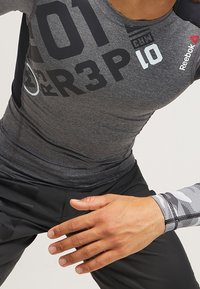 Šedé sportovní tričko s černými akcenty a tištěnými čísly, doplněné černými kalhotami a vzorovaným rukávem. Materiál vypadá hladce a je přiléhavý.