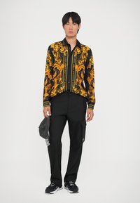 Camicia nera e oro con un design intricato, abbinata a pantaloni cargo neri e sneakers nere. Mano che tiene un cappellino grigio.