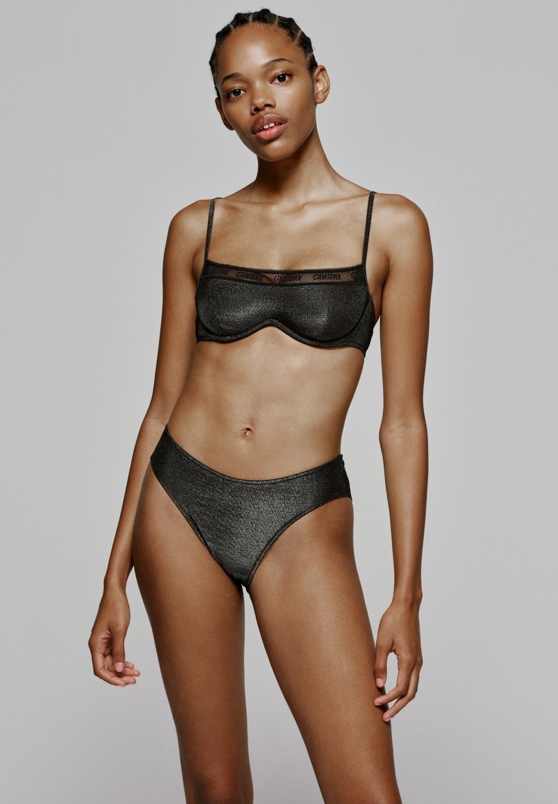 Zwarte bh en hoge bikiniset in een gestructureerde, metallic stof. De bh heeft smalle bandjes en een semi-doorzichtige rand aan de bovenkant.