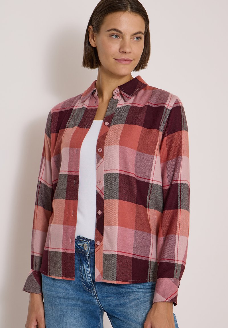 Femme portant une chemise à carreaux rouge, rose et grise par-dessus un haut blanc et un jean bleu, regardant légèrement sur le côté.