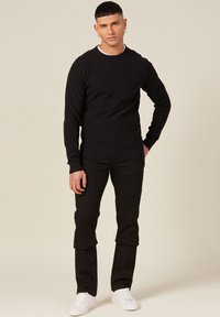 Schwarzer Strickpullover mit geripptem Muster, kombiniert mit schwarzen Hosen und weißen Sneakern. Einfaches und schlankes Design mit Rundhalsausschnitt.