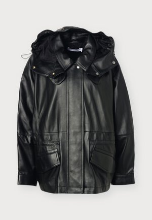 Persona que lleva una chaqueta de cuero negra suelta con cuello grande y pantalones negros de pierna ancha, de pie frente a un fondo claro y liso.