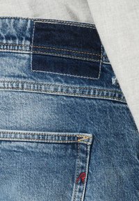 Tasca dei jeans in denim blu con doppie cuciture, una toppa in denim scuro sopra e un piccolo dettaglio ricamato rosso nell'angolo della tasca.