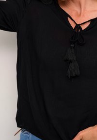 Svart blus med knytkrage och två tofsar. Har ett lätt, strukturerat tyg. Inkluderar dekorativa detaljer på axlarna.