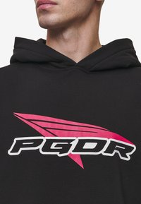 Černá mikina s kapucí s růžovo-bílým grafickým logem obsahujícím "PGDR" a stylizovaný design. Materiál působí měkce s texturovaným povrchem.