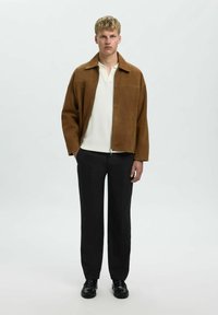 Giacca in suede marrone con zip frontale, colletto a punta e due tasche sul petto, abbinata a pantaloni neri e scarpe nere.