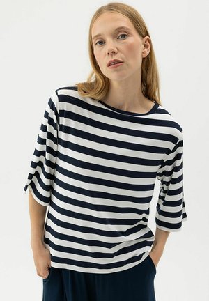 Heledate juustega naine, kes kannab lahtist musta-valge horisontaaltriibulist särk ja tumedaid pükse, käed taskutes, taustal lihtne taust.