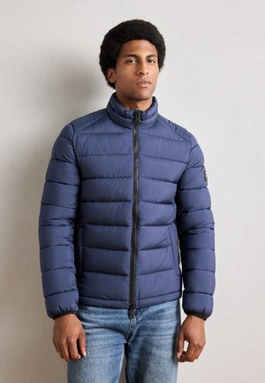 Navy blue puffer jacket with horizontal quilted sections, high collar, zipper front closure, and ribbed cuffs, paired with blue jeans.
