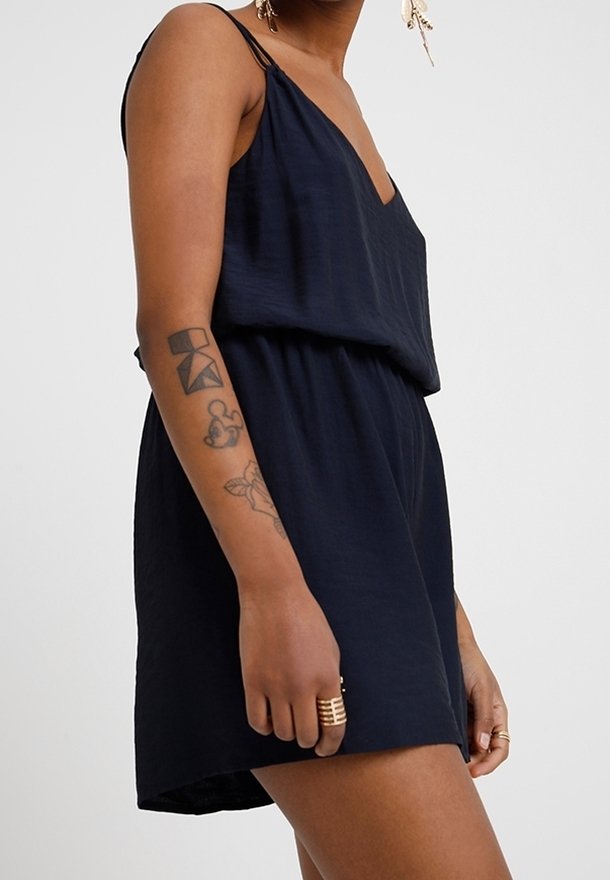 Granatowy kombinezon na cienkich ramiączkach, z głębokim dekoltem w kształcie litery V, luźnym fasonem tułowia i elastycznym paskiem w talii; wykonany z lekkiego materiału, charakteryzujący się swobodnym krojem.