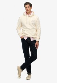 Crème hoodie met kangoeroezak en lichtgrijze tekst, gelaagd over een roze shirt, gecombineerd met een donkere spijkerbroek en beige sneakers.