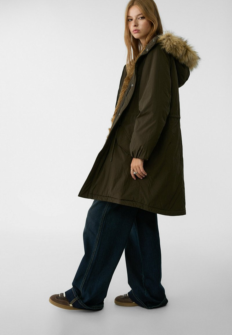 Parka longue vert olive avec une capuche doublée de fourrure, dotée d'une fermeture éclair et de boutons-pression, de poches latérales et d'une coupe décontractée sur un jean en denim ample.