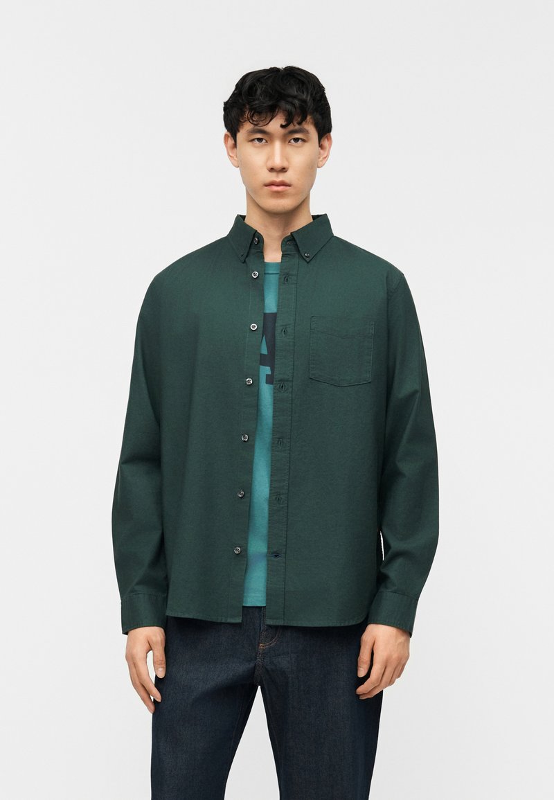 Νεαρός άνδρας που φοράει σκούρο πράσινο πουκάμισο με κουμπιά πάνω από μια τιρκουάζ μπλούζα με γραφικό σχέδιο και σκούρο μπλε τζιν, στέκεται μπροστά σε ένα απλό ανοιχτόχρωμο φόντο.