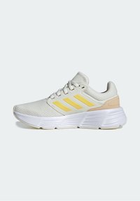 Scarpa da ginnastica bianca con tomaia in rete, strisce gialle e accentuazione del tallone beige. Presenta un design testurizzato e una suola ammortizzata per il comfort.