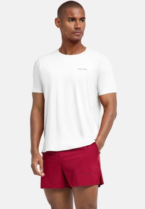 Mężczyzna w prostym białym T-shircie z krótkim rękawem i czerwonych sportowych szortach stoi z jedną ręką w kieszeni na jasnym tle.