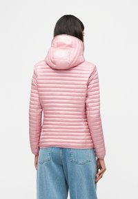 Γυναίκα που φοράει ένα ανοιχτό ροζ καπιτονέ μπουφάν με κουκούλα, συνδυασμένο με άνετο ανοιχτό μπλε τζιν παντελόνι, που στέκεται γυρισμένη προς τα πίσω με φόντο λευκό.