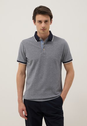 Homme portant un polo gris à manches courtes avec col et bordure bleu marine, debout avec une main dans la poche sur un fond uni.