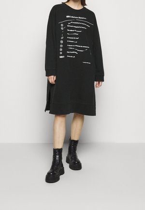 Personne portant une robe midi noire à manches longues avec un texte blanc imprimé et des bottines à plateforme noires à lacets, debout sur un sol gris.