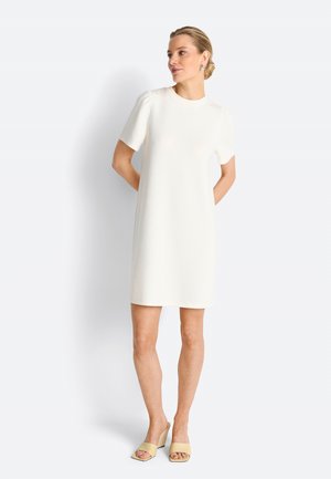 Donna in piedi, indossa un semplice vestito bianco a maniche corte fino al ginocchio e sandali beige con tacco, con le mani dietro la schiena.