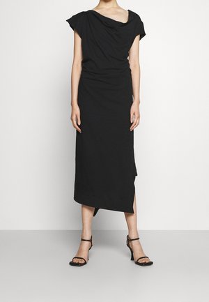 Vrouw met een zwarte, asymmetrische midi-jurk met mouwloze capes en zwarte hoge hakken met enkelbandjes, staand tegen een witte achtergrond.