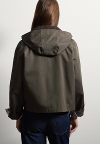 Giacca con cappuccio verde oliva dal texture morbido, con colletto in corduroy marrone e polsini a motivo scozzese. Realizzata in materiale leggero.