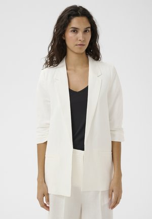 Kobieta o długich kręconych włosach, ubrana w biały żakiet i spodnie, na czarnej bluzce z dekoltem w serek, stojąca na jednolitym jasnym tle.