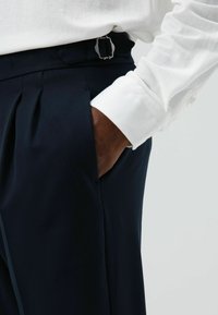 Pantalon plissé marine avec une boucle argentée à la taille, associé à une chemise blanche texturée à manches longues. Finition en tissu satinée.