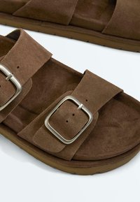 Bruine suède sandalen met twee verstelbare zilveren gespen en een gevoerde binnenzool op een lichte achtergrond.