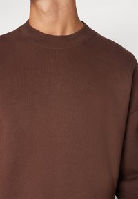 Sudadera de cuello redondo marrón, confeccionada en una tela suave, con un corte holgado y un cuello ribeteado. Sin patrones ni acentos visibles.