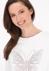 Felpa bianca con un design di farfalla in strass colorati sulla parte anteriore. Il tessuto ha una texture morbida con un colletto a coste.