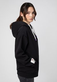 Zwarte rits-hoodie met een trekkoordkap, voorzien van een bloemenaccent op de manchetten en een voorzak. Glad materiaal, losse pasvorm.