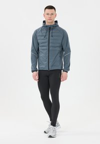 Mand står iført en blå polstret hættetrøje, sorte leggings og hvide løbesko på en hvid baggrund.