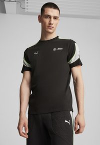 Musta urheilu t-paita, jossa on vihreitä ja vaaleanharmaita yksityiskohtia, ja jossa on Mercedeksen ja Puman logot. Materiaali näyttää sileältä ja sovituu hyvin vartaloon.