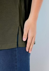 Olivengrüne Baumwoll-T-Shirt mit seitlichen Schlitz, mit abgerundetem Saum. Hand liegt auf dem Shirt in der Nähe der Naht gegen blaue Denim-Jeans.