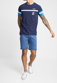 Marinblå t-shirt med vita och blå ränder, med texten 'CORE' och '1990'; tillsammans med blå shorts och vita sneakers. Bärs av en person.