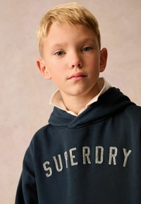 Sudadera con capucha de color azul marino con "SUPERDRY" bordado en gris claro. Tela suave, capucha amplia, cuello en capas, con un ajuste relajado.