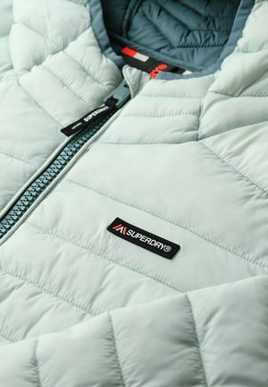 Giacca imbottita azzurro chiaro con texture trapuntata, caratterizzata da una chiusura frontale con zip e un'etichetta nera con logo "Superdry" vicino al collo.