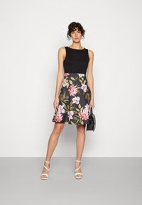 Black sleeveless top with a floral-patterned A-line skirt in dark background colors, paired with white strappy sandals and a small black handbag.