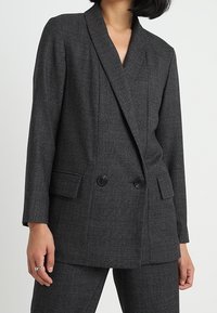 Šedý kostkovaný dvouřadý blazer s klopovým límcem, dvěma předními kapsami a černými knoflíky. Hladká textura a přizpůsobený design.
