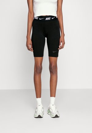 Osoba nosząca czarne rowerowe spodenki marki Nike, biały krótki top, białe skarpetki Nike i białe trampki, stojąca na jednolitym tle.