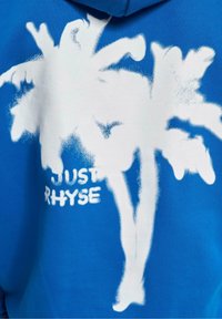 Tissu bleu avec des palmiers blancs peints à la bombe et texte partiellement visible indiquant "JUST RHYSE."