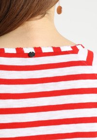 Rot-weiß gestreifes Oberteil mit rundem Ausschnitt, ausgestattet mit einem kleinen schwarzen Knopf auf der Rückseite. Weicher Stoff, lässiges Design.