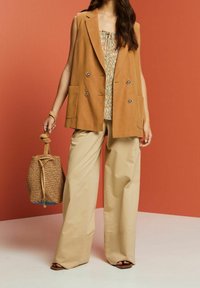 Femme portant un gilet sans manches brun, une blouse imprimée, un pantalon large beige, tenant un sac en osier, se tenant devant un fond orange.