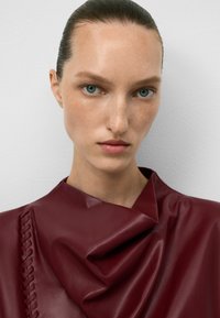 Indumento in pelle burgundy con design drappeggiato e dettagli testurizzati. Presenta un accento intrecciato sul lato sinistro, superficie liscia e una leggera lucentezza.